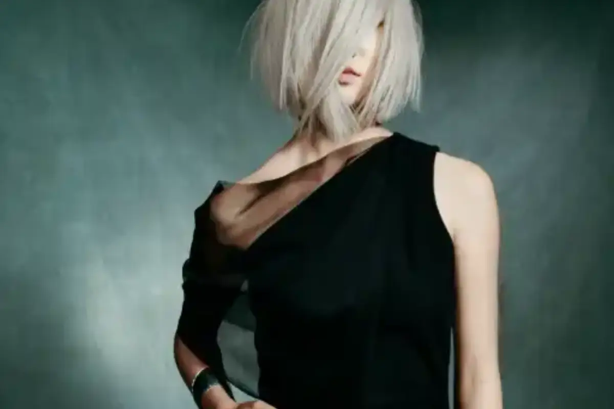 Person with a chin-length platinum blonde bob, styled with aveda-hair-care, wears a one-shoulder black dress, standing against a dark, blurred background. - Jax Salon and Spa