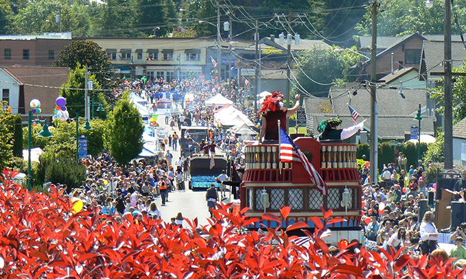 A crowded street parade features decorated floats, people waving American flags, and spectators lined up along the sidewalks on a sunny day. - Jax Salon and Spa