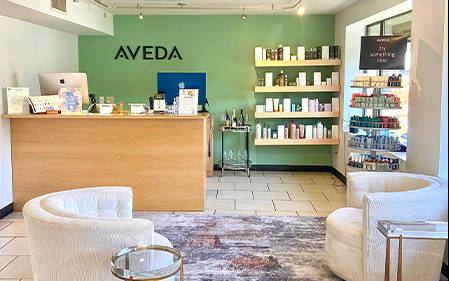 Reception area of an Aveda salon with a wooden desk, product shelves on a green wall, two white chairs, and a rug on a tiled floor. - Jax Salon and Spa