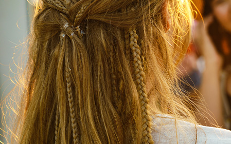 A person with long, blonde hair styled in multiple small braids and loose strands, photographed from behind in natural sunlight. - Jax Salon and Spa