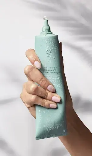 A hand holds a light blue-green tube with a droplet of gel at the tip, set against a soft, radiant background—perfect for those looking to enhance existing color with demi color brilliance. - Jax Salon and Spa