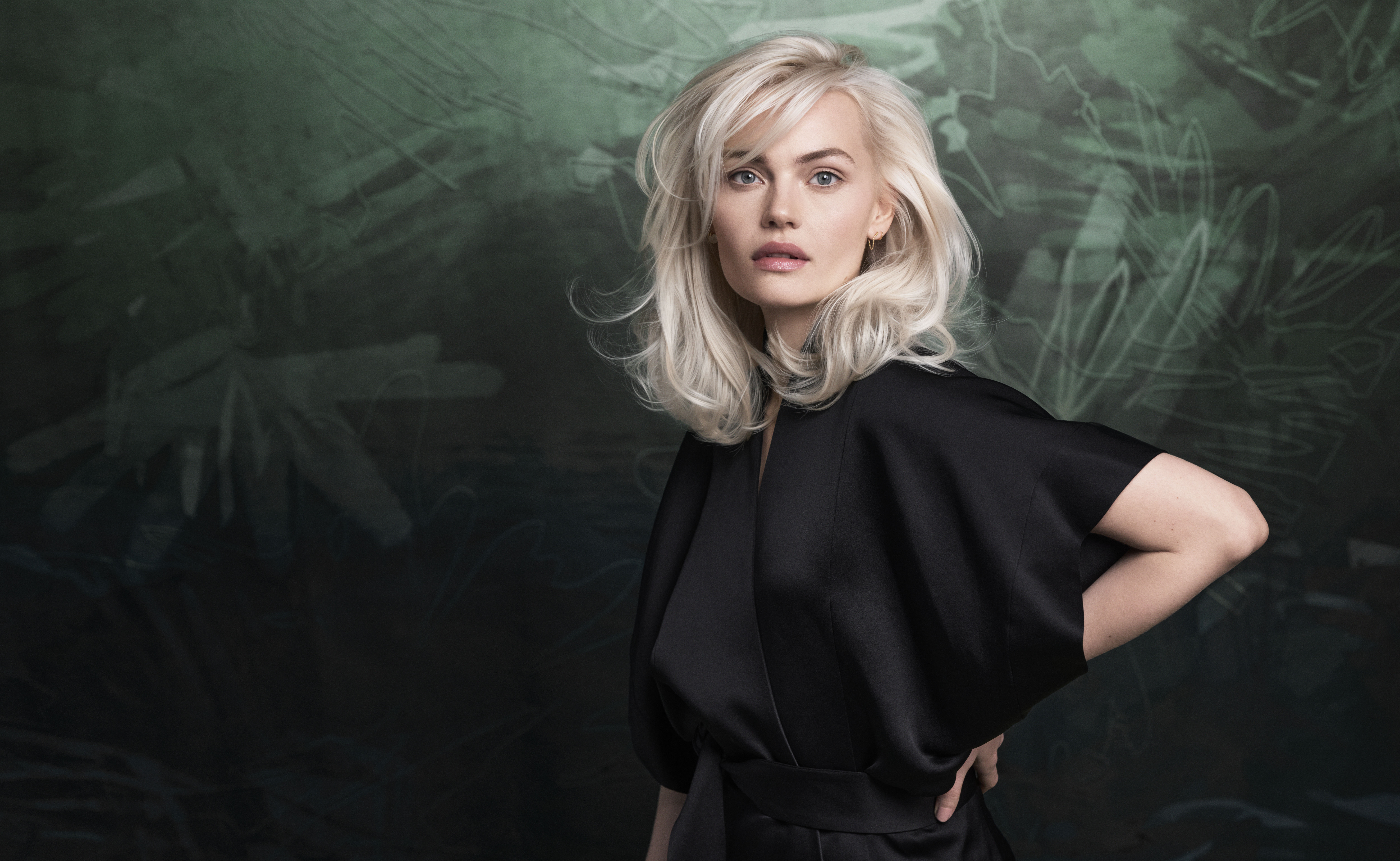 A woman with platinum blonde hair wearing a black robe stands in front of a dark green abstract background, looking at the camera. - Jax Salon and Spa