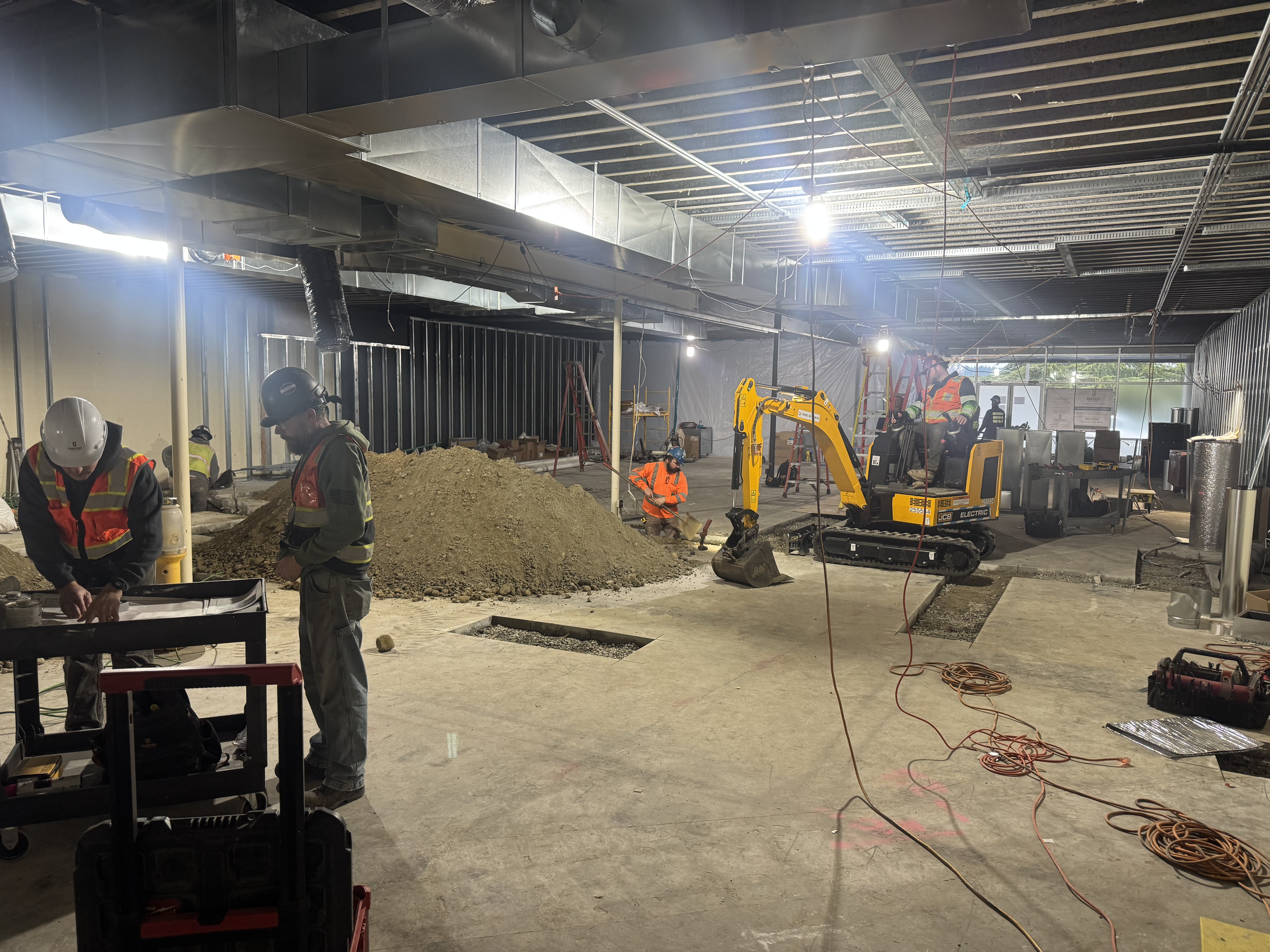 Construction workers with safety gear operate equipment and work inside a partially finished building with exposed ceilings, soil piles, and a small excavator. - Jax Salon and Spa