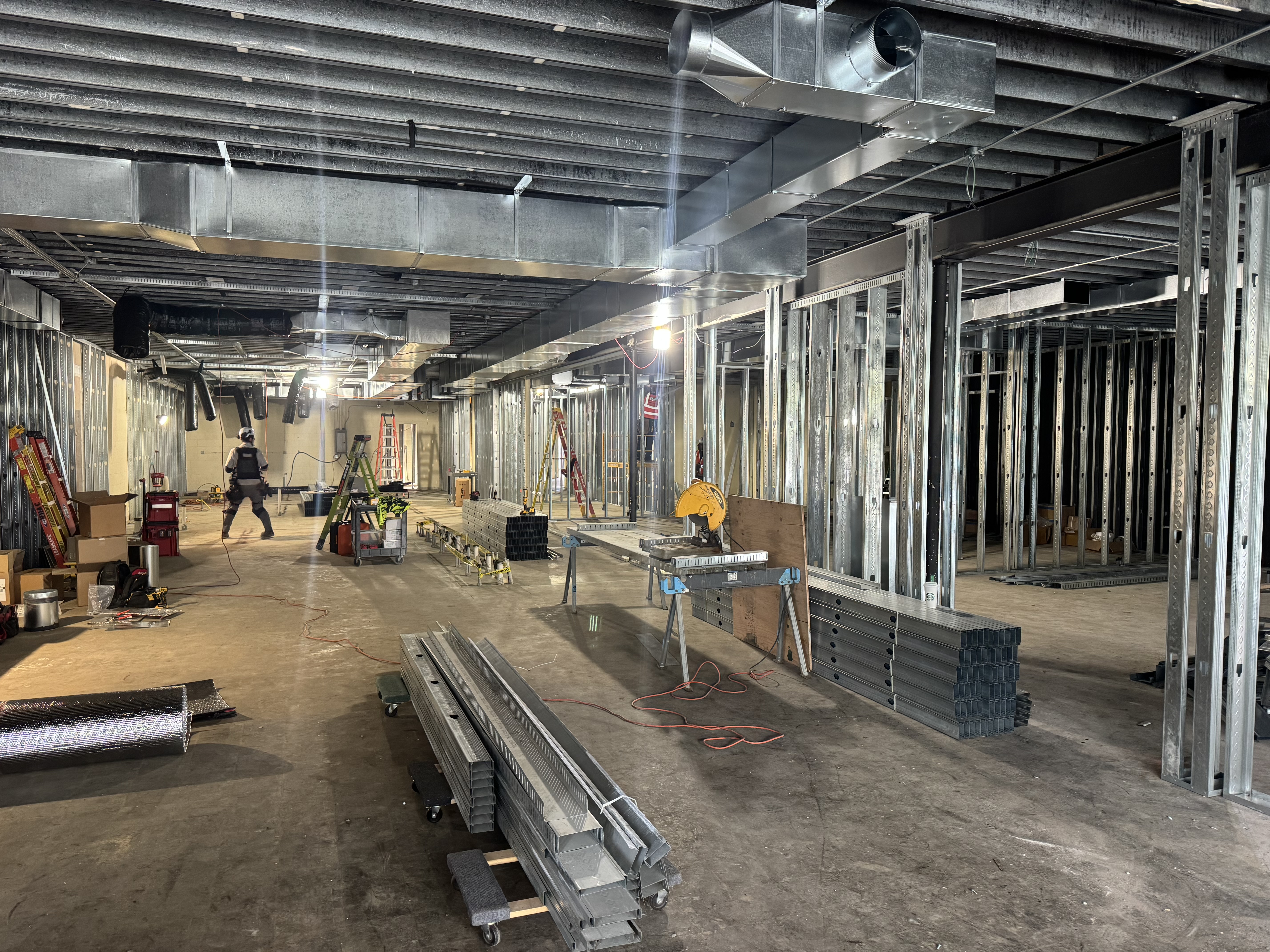 Construction site with exposed metal wall framing, ductwork, tools, and building materials; one worker is present in the background. - Jax Salon and Spa