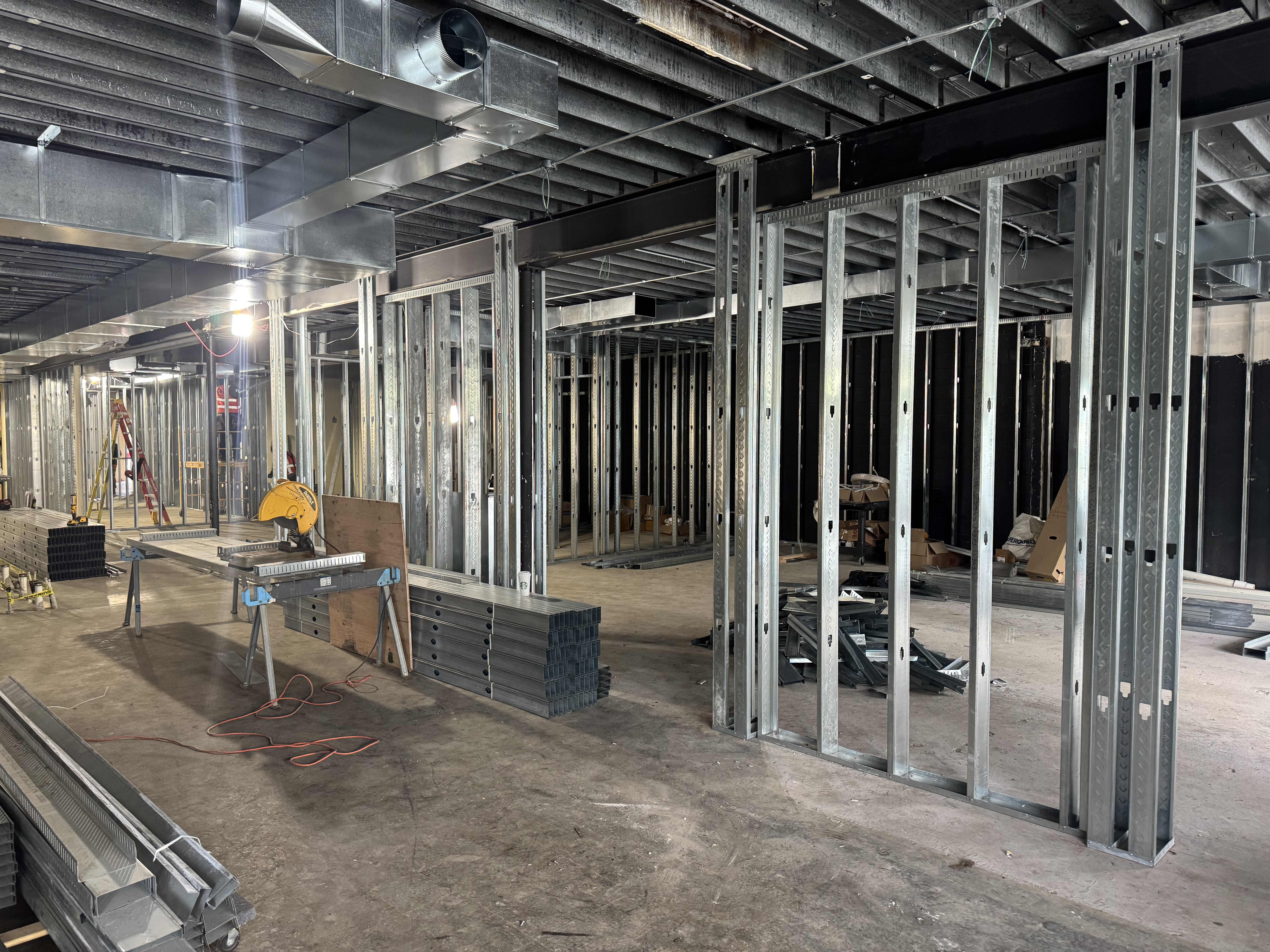 Interior construction site showing metal wall studs, exposed ceiling beams, ductwork, and various construction materials and tools scattered on the unfinished floor. - Jax Salon and Spa