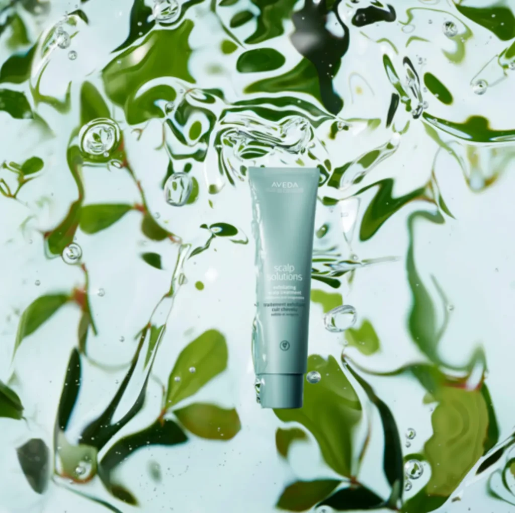 A tube of Aveda Scalp Solutions Exfoliating Scalp Treatment is suspended in water with green leaves visible in the background. - Jax Salon and Spa