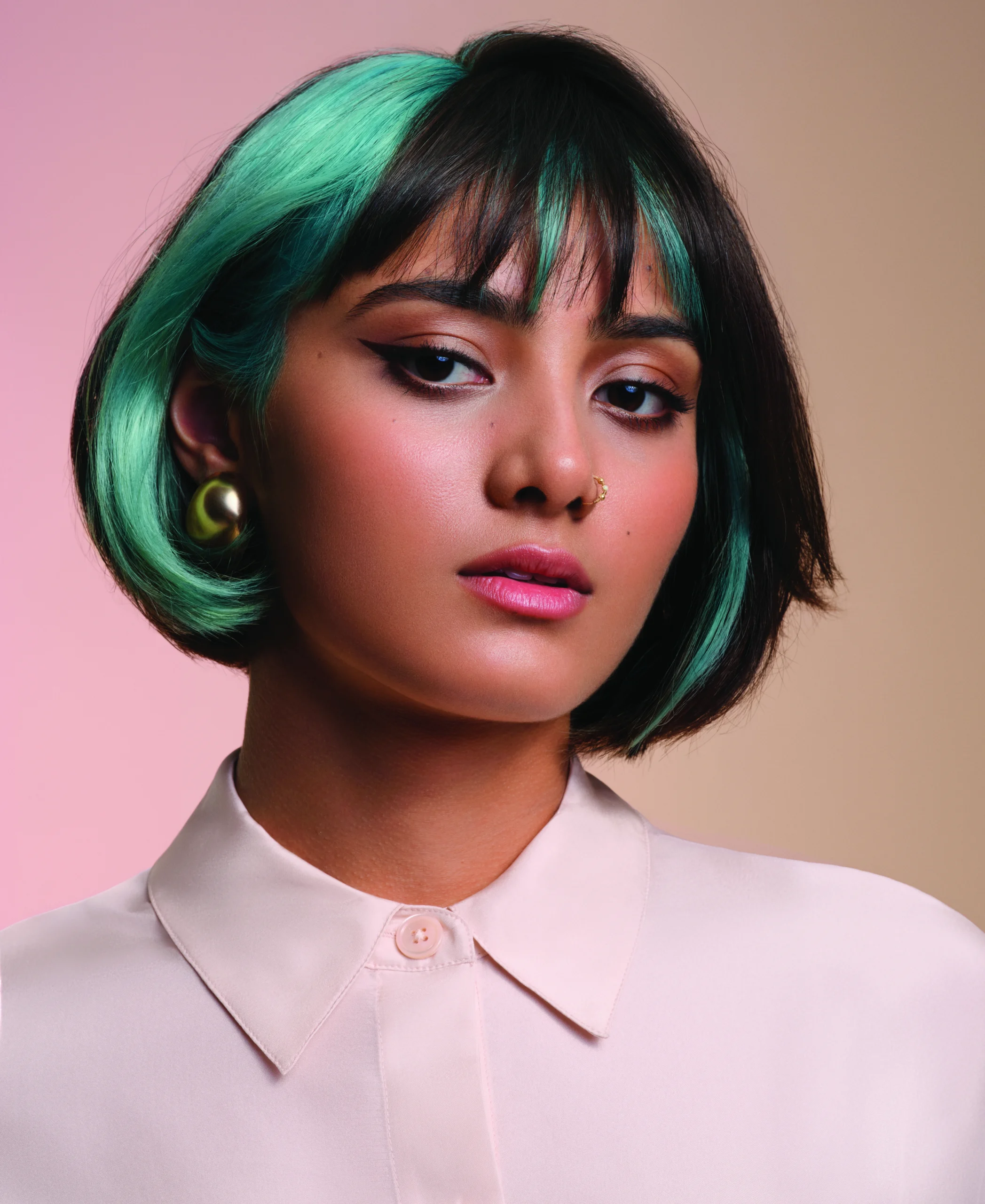 A person with short black hair featuring green highlights, wearing a white collared shirt, gold earrings, and a nose ring, poses against a pastel background. - Jax Salon and Spa