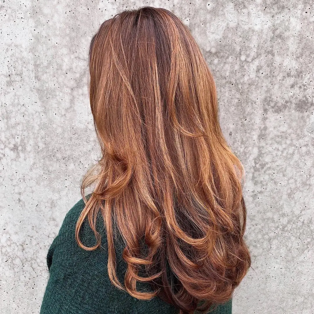 Person with long, wavy light brown hair wearing a dark green sweater, standing with their back to the camera in front of a textured gray wall. - Jax Salon and Spa