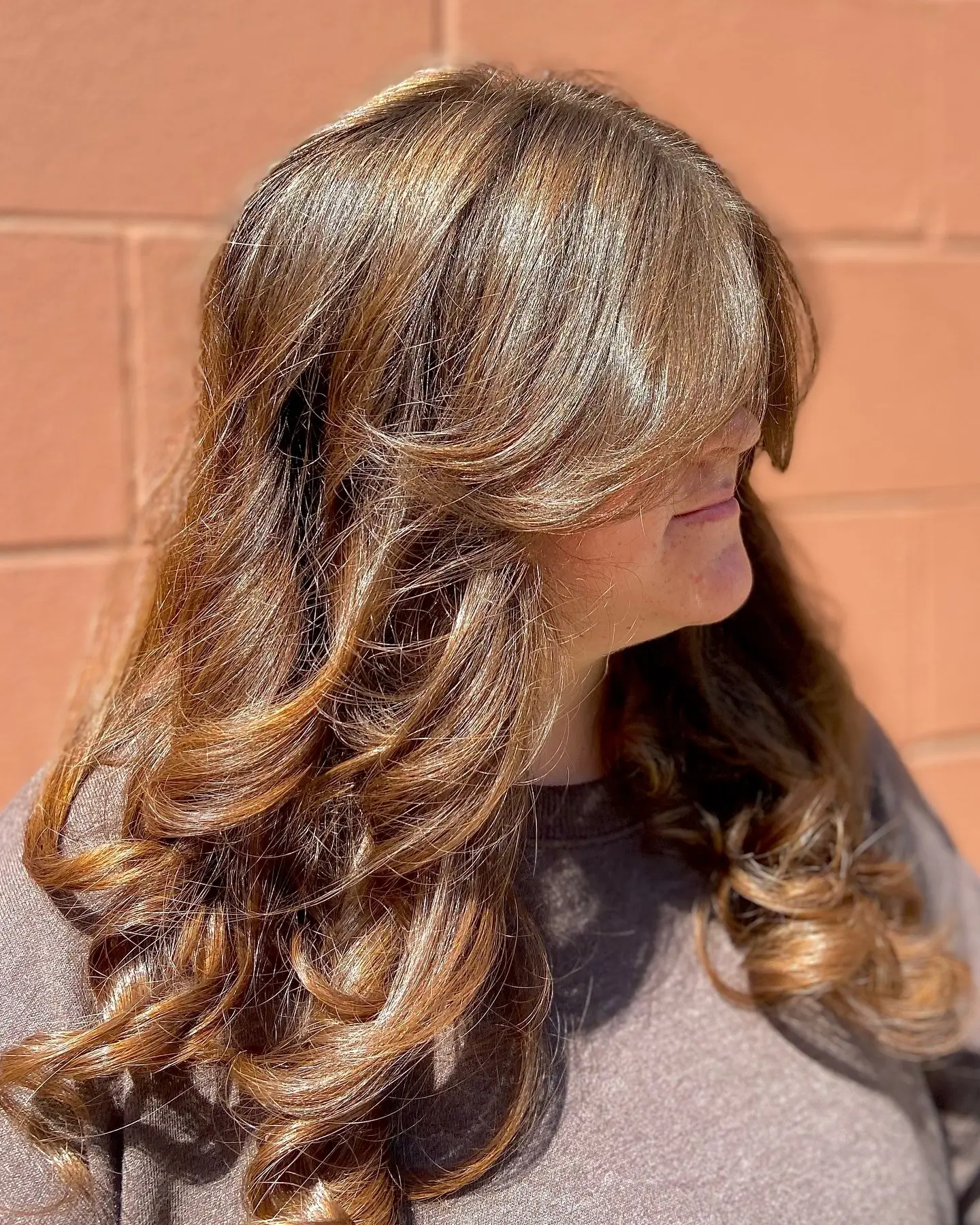 A person with long, wavy, light brown hair styled in voluminous layers and bangs stands in front of an orange brick wall. - Jax Salon and Spa