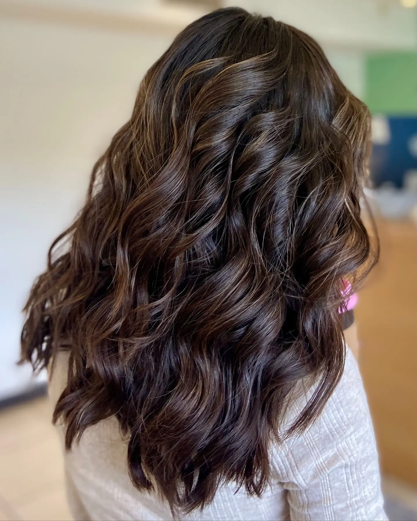 Person with long, wavy, dark brown hair viewed from the back. The hair has soft curls and subtle highlights. The person is wearing a light-colored top. - Jax Salon and Spa