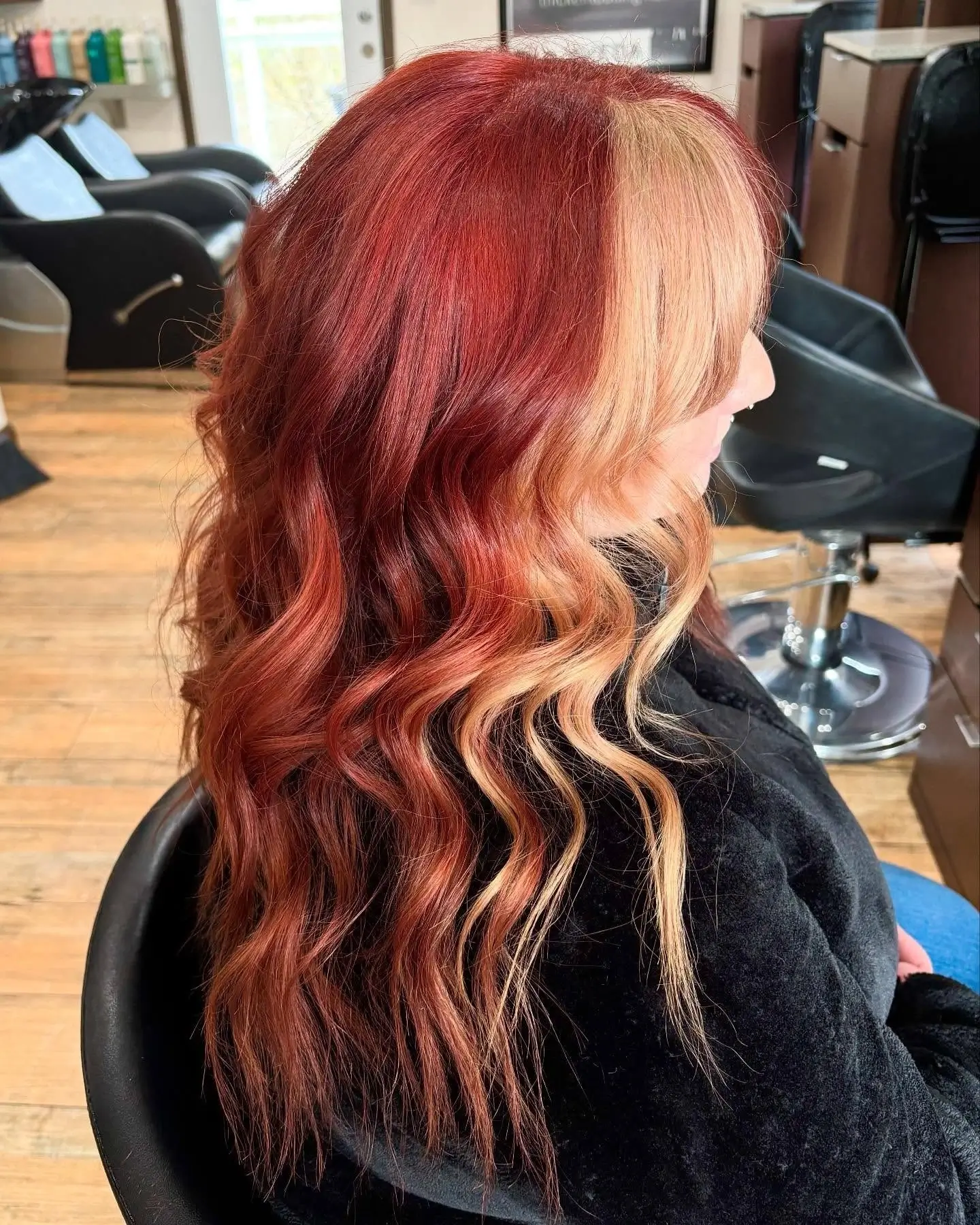 Person with long, wavy hair dyed in vibrant red with blonde highlights sits in a salon chair, facing right. - Jax Salon and Spa