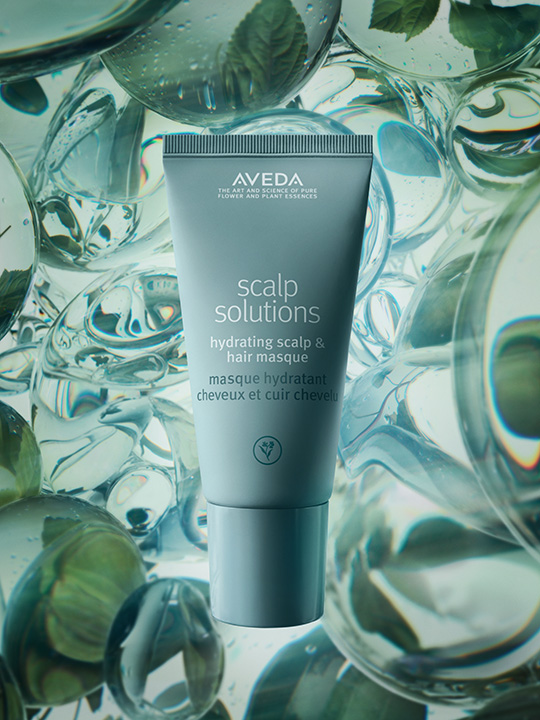 A tube of Aveda Scalp Solutions Hydrating Scalp & Hair Masque stands upright against a background of transparent globules and green leaves, highlighting its dedication to scalp care and promoting optimal scalp health. - Jax Salon and Spa