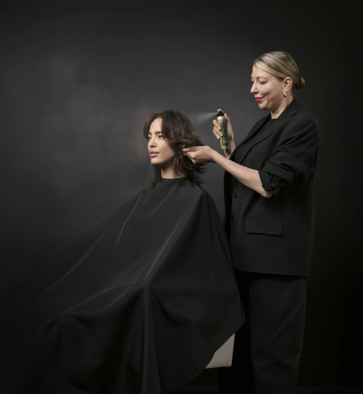 A hairstylist wearing black sprays a mist onto a seated woman's hair, who is covered with a black cape, against a dark background. - Jax Salon and Spa