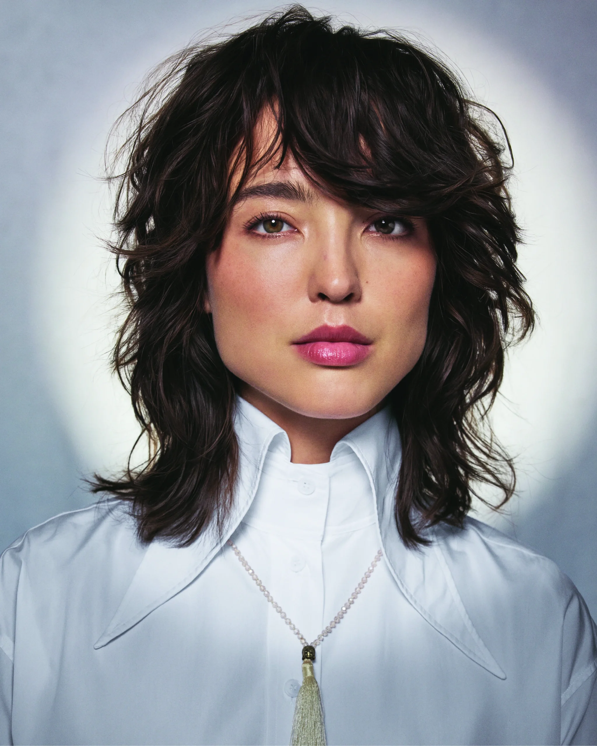 A person with wavy, layered brown hair and a white collared shirt with a tassel necklace, looking directly at the camera against a neutral background. - Jax Salon and Spa