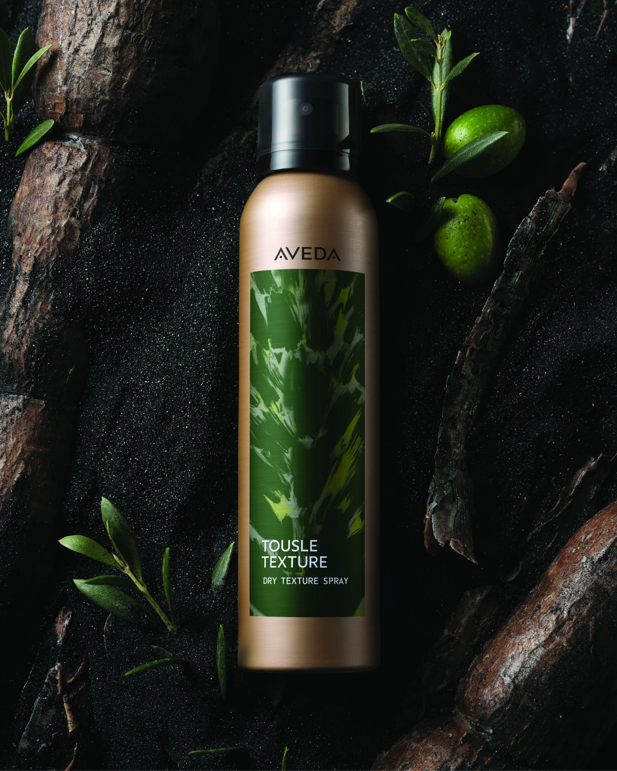 Aveda Tousle Texture Dry Texture Spray canister placed on a dark surface with green leaves and small green fruits around it. - Jax Salon and Spa