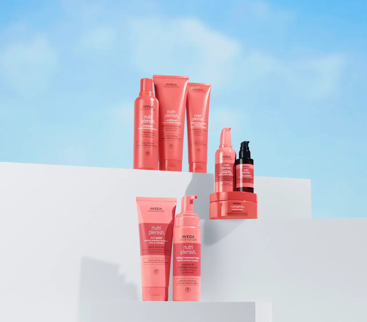 Multiple Aveda Nutri Plenish hair care products, including their frizz control spray, are displayed in coral-colored bottles on white stairs against a blue sky background. - Jax Salon and Spa