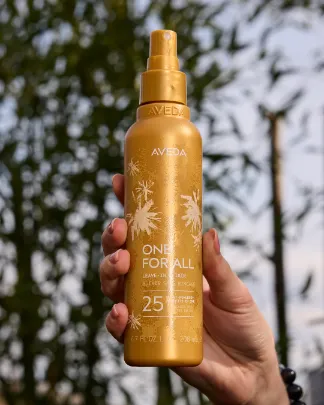 A hand holds a gold bottle of Aveda One For All 25 Benefits Leave-In Spray against a blurred outdoor background with green plants. - Jax Salon and Spa