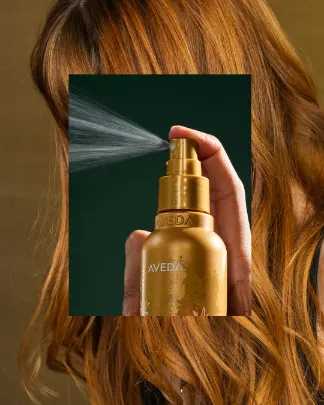 A close-up of a hand spraying a golden Aveda bottle, overlaid on long, wavy, reddish-brown hair in the background. - Jax Salon and Spa