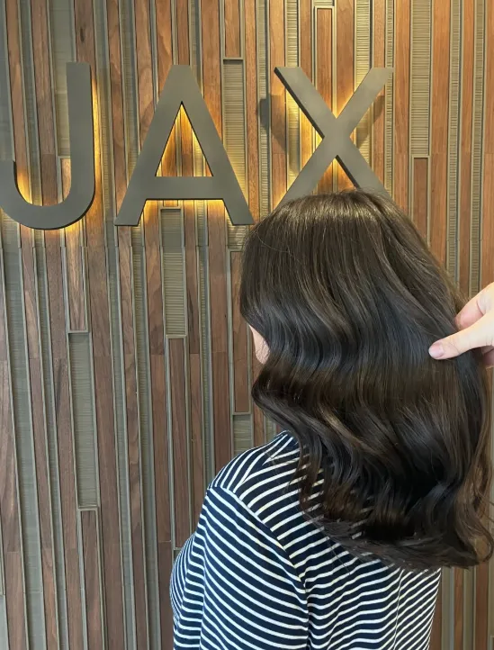 A person with shoulder-length, wavy dark brown hair styled for Halloween, standing in front of a wooden wall with large letters spelling “JAX.” - Jax Salon and Spa